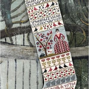 May include: A cream-colored fabric Christmas stocking featuring intricate cross-stitch embroidery. The design includes rows of birds, floral motifs, alphabet letters, and a central figure in a red dress. Text reads: It is a lovely thing for youth to hate a lie and speak the truth and Let truth & virtue be your guide and wisdom over your ways preside.