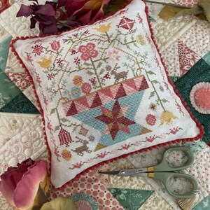 May include: A white pillow with a red, pink, and blue embroidered design of a flower pot with flowers and a deer. The pillow has a red pom-pom trim.