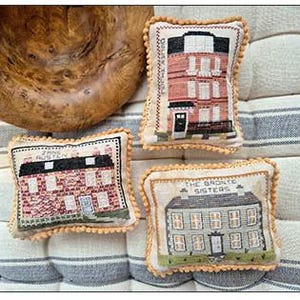 May include: Three decorative rectangular pillows with embroidered house designs. The pillows feature cream-colored fabric with a pom-pom trim. The houses are depicted in various colors, including red, black, and white, with the text "Jane Austen" and "The Bronte Sisters".