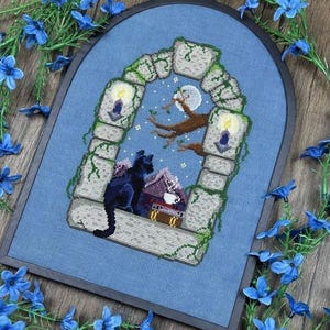 May include: Cross-stitch art featuring a black cat perched in a stone archway, gazing at a moonlit night scene. The design includes a house, candles, and a tree branch. The artwork is framed in a blue fabric.