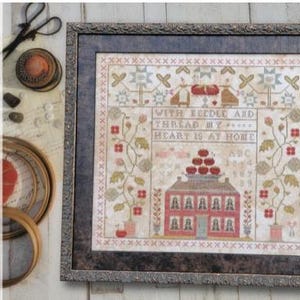 May include: Framed cross-stitch sampler with a house, floral elements, and the text "WITH NEEDLE & THREAD MY HEART IS AT HOME." The sampler is in a dark gray frame. Additional sewing tools are visible.