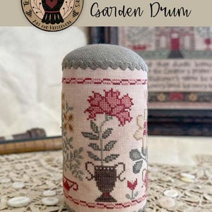 May include: A cylindrical pincushion featuring cross-stitch embroidery on light pink fabric. The design displays a dark brown urn holding a large red flower, surrounded by smaller floral motifs. It has grey scalloped trim at the top and bottom edges.