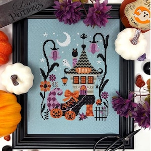 May include: A framed cross-stitch embroidery on light blue fabric featuring a black shoe-shaped house with a pointed roof, the text BOO, a crescent moon, stars, a ghost, a black cat, and two jack-o'-lanterns, surrounded by purple flowers.