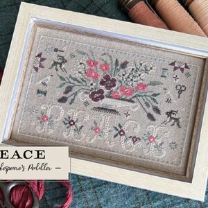 May include: Framed cross-stitch art with a floral design in pink, burgundy, and white. The word "PEACE" is stitched below the flowers. The frame is light-colored wood. The text "PEACE - by Shakespeare's Peddler" is in a rectangular label.