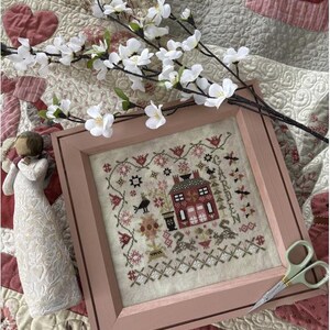 May include: A framed cross-stitch embroidery with a pink frame. The embroidery depicts a small red house with a white picket fence and a garden with flowers and a bird. The text "GARDEN" is stitched in the upper right corner.