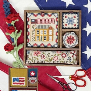 May include: A patriotic-themed craft box with cross-stitch designs, including an American flag, a house, and floral patterns. The box also contains a pair of red-handled scissors and a crochet flower arrangement. The text "Patriotic Box" is visible.