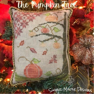 May include: A cross-stitch pillow featuring a whimsical pumpkin tree with orange pumpkins, green leaves, and red and brown leaves falling from the branches. The pillow has a gold trim and the text "The Pumpkin Tree" at the top.