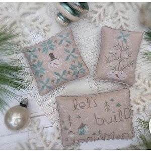 May include: Three beige cross-stitch pillows with winter-themed designs. One features a snowman, another a tree, and the third says "let's build a snowman." The image includes ornaments, pine needles, and snowflakes, creating a festive holiday scene.