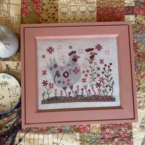 May include: A square cross-stitch embroidery featuring a light blue hen and a smaller bird surrounded by pink and dark red floral motifs on a white fabric background, encased in a solid dusty pink wooden frame with a flat, wide border design.