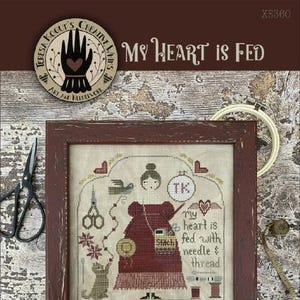 May include: Cross stitch pattern featuring a woman with a needle and thread, surrounded by sewing supplies. The pattern is titled "My Heart is Fed" and is a Nashville Exclusive by Teresa Koour's Creative Labs.