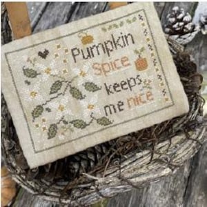 May include: Cross-stitch sampler with the text "Pumpkin Spice keeps me nice" in brown and green. The sampler features floral and pumpkin motifs, set against a beige background. The sampler is displayed in a woven basket with pinecones.