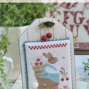 May include: A cross-stitch design titled "CHERRY ON TOP" by Brenda Gervais. The design features a bunny holding a basket of flowers and cherries. The design is framed with a wooden plaque and adorned with felt cherries and leaves.