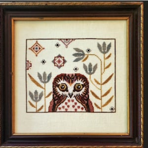 May include: Cross-stitch artwork of an owl with large yellow eyes, framed in a dark wood frame. The owl is centered against a cream-colored background with floral and geometric patterns in brown, gray, and red. The frame is approximately 10 inches square.