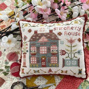 May include: A decorative, rectangular pillow with a cross-stitch design featuring a house and the words "stitchery house." The pillow has a white background with a red border and is surrounded by flowers.