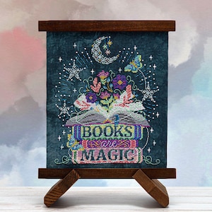 May include: A framed cross-stitch piece on a wooden easel. The artwork features a stack of books with the words "BOOKS are MAGIC" stitched in colorful thread. Above the books are embroidered flowers, a crescent moon, stars, and a butterfly, all against a dark teal background.