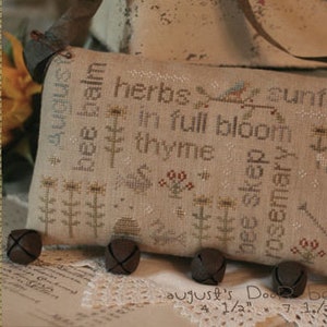 May include: A beige fabric pillow with a cross-stitch design featuring herbs, bees, and a sunflower. The text on the pillow reads "bee balm, herbs in full bloom, thyme, bee skep, rosemary". The pillow is 4 1/2 inches by 7 1/2 inches.