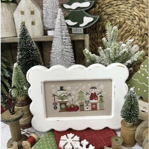 May include: A framed cross-stitch featuring snowmen and Christmas trees. The frame is white with a scalloped edge. The image is surrounded by miniature Christmas trees, wooden spools, fabric, and decorative snowflakes. Text on the image reads "Snowman 2025" and "Primrose Cottage."