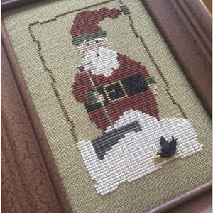 May include: A framed cross-stitch design featuring a red and white Santa Claus holding a green and white candy cane. The Santa is standing on a white snowdrift with a black bird perched on the edge.