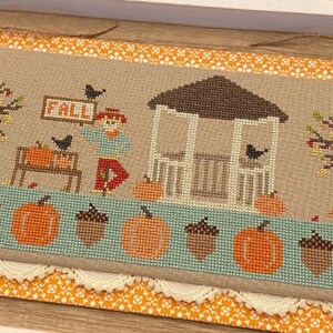 May include: Cross-stitch design featuring a fall scene with a gazebo, pumpkins, and a scarecrow holding a "FALL" sign. The design includes trees with autumn leaves and a border of pumpkins and acorns. The color palette includes orange, brown, and green.