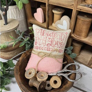 May include: A pink and cream-colored decorative pillow with the text "Abide In My Love" is tied with twine and placed in a wooden bowl. The image includes wooden spools, scissors, and the text "Primrose Cottage".