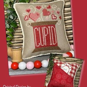 May include: A decorative pillow with a cross-stitched design featuring the word "CUPID" in red, with hearts above. A smaller pillow with a red pattern and a gingham pocket is also shown. The design is by Crafty Bluebonnet Designs.