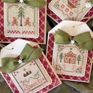 May include: Five handmade Christmas ornaments with a winter theme. Each ornament features a different design, including a gingerbread house, a snowy forest, and a cabin. The ornaments are made with fabric and embroidery and are decorated with green ribbon and white snowflakes.