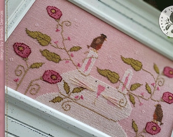 May include: Cross-stitch design titled "Spring Fling" featuring two white rabbits, pink flowers, and brown birds. The design is framed in a white wooden frame. The text "SPRING FLING" and the designer's name are visible.
