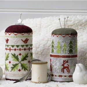 May include: Two cylindrical pincushions with cross-stitch designs. One has a burgundy top and a white base with green and red accents. The other has a green top and a gray base with green trees and a red reindeer. A spool of thread and scissors are also present.