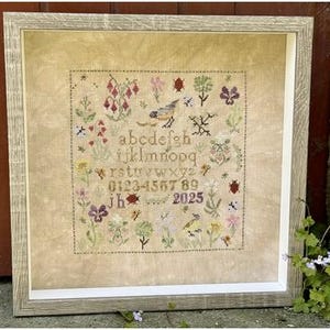 May include: Framed cross-stitch sampler featuring a floral and bird design with the alphabet, numbers, and the year 2025. The artwork is in a light-colored frame. The sampler includes various flowers, birds, and insects in shades of green, purple, and red.