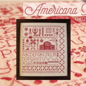 May include: A framed cross-stitch sampler with a red and white color scheme. The sampler features a variety of designs, including a house, a pumpkin, a flower, and a heart. The alphabet is also included in the design. The sampler is titled "Americana Red" and has the product code "OH1256".