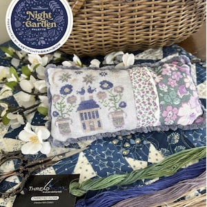 May include: A cross-stitch pillow with a night garden design featuring a house, flowers, and birds. The pillow is on a blue and white quilt with a basket and embroidery floss.