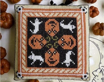 May include: A square, cross-stitched Halloween decoration featuring orange pumpkins and white ghosts on a black background. The design is framed with a cream and orange border, with small pumpkins scattered around the edges.