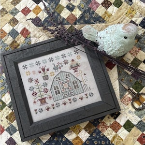 May include: A framed cross-stitch sampler with a farmhouse design in a dark gray frame. The sampler features a greenhouse, flowers, and a rooster. A decorative bird and lavender sprigs are nearby, set on a patchwork quilt.