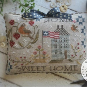 May include: A cross-stitch pillow with a white background and a red, white, and blue house with an American flag. The pillow also features a robin, strawberries, and the text "Sweet Home".