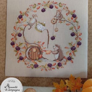 May include: A cross-stitch embroidery on light beige fabric featuring a circular wreath of autumn leaves and dark purple blackberries. Inside the wreath, two mice and a hedgehog are depicted near a small round door, with one mouse pouring tea.