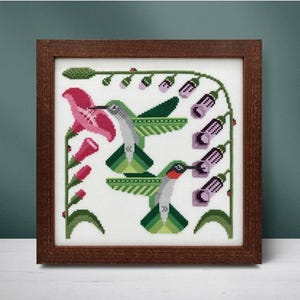 May include: Cross-stitch art of two hummingbirds with green and gray bodies, red throats, and outstretched wings, feeding from pink and purple flowers. The artwork is framed in a dark brown wooden frame.