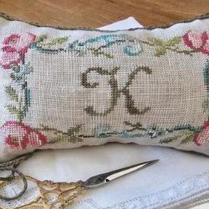 May include: A rectangular, off-white cross-stitch pillow with a floral border and the letter "K" in the center. The flowers are pink and the border is green and blue. A pair of gold scissors and a decorative cord are in the foreground.