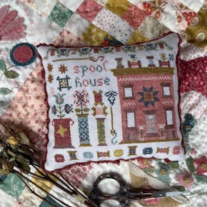 May include: A square decorative pillow featuring a cross-stitch pattern on white fabric. The design includes the text "spool house" in dark letters, a multi-story building, various colorful thread spools, and floral motifs in red, blue, and green.