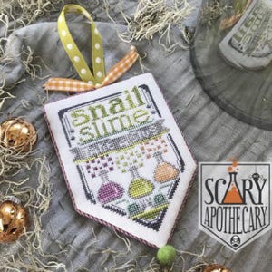 May include: A cross-stitched banner with the words "Snail Slime" in green and gray. The banner features colorful potion bottles and is adorned with a yellow polka dot ribbon and a green pom-pom. The Scary Apothecary logo is visible.