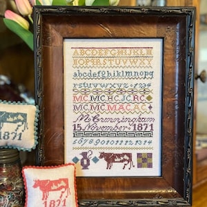 May include: A framed cross-stitch sampler with a white background and colorful embroidery. The sampler includes the alphabet, numbers, and the text "Mo Cunningham 15 November 1871".