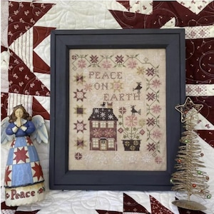 May include: A framed cross-stitch sampler with a white background and colorful embroidery. The sampler features a house with a red roof, a tree with pink flowers, and the text "Peace on Earth".