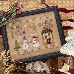 May include: Framed cross-stitch artwork with a winter theme. The design features two snowmen, a house, a Christmas tree, and the text "I SMELL SNOW". The frame is black and ornate, with a beige background.