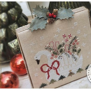 May include: A cross-stitch pattern featuring a white swan with a red bow, surrounded by greenery and red berries. The pattern is framed and displayed on a beige background with a snowflake pattern. The text "Christmas Swan" is written in cursive on the left side of the image. The text "design by Brenda Gervais" is written in cursive below the text "Christmas Swan". The text "#CS352" is written in the top right corner of the image.