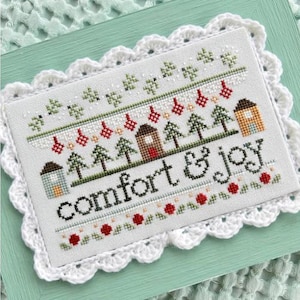 May include: A cross-stitch design with the words "comfort & joy" in black thread. The design features small houses, Christmas trees, and red ornaments. The piece is framed with a white, scalloped lace border and set on a light green background.