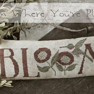 May include: A rectangular, off-white decorative pillow with the words "Bloom Where You're Planted" embroidered in brown. The design includes flowers and a hummingbird. The pillow is suspended by a twine loop and surrounded by greenery.