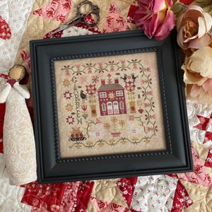 May include: Framed cross-stitch artwork featuring a red house with floral and bird motifs. The artwork is in a black frame and includes the word "GARDEN". The color palette includes red, pink, and beige tones.
