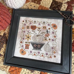 May include: Framed cross-stitch art featuring a fall harvest theme. The design includes pumpkins, flowers, and a witch. The frame is dark gray. The artwork is displayed on a patterned quilt.