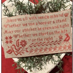 May include: A cross-stitched sampler with red embroidery on a tan background, featuring the text "Bless us all with needle in hand." Decorative floral and bird motifs border the text. The sampler is set in a heart-shaped dish with greenery.