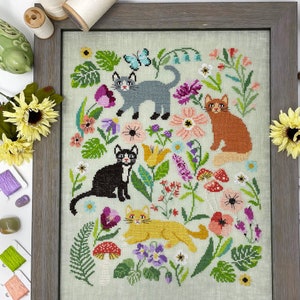 May include: Cross-stitch embroidery featuring four cats in a garden setting. The cats are surrounded by colorful flowers, leaves, and mushrooms. The cats are black and white, gray, orange, and yellow.
