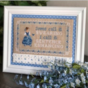 May include: Cross-stitch embroidery framed in white with a beige background. The design features a girl in a blue dress holding a bird with the text "Some call it swearing I call it sentence enhancing".